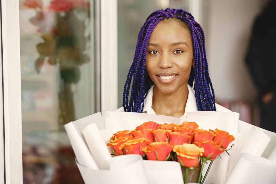 A young woman with dark skin and long, twisted purple braids is smiling as she holds a large bouquet of fresh, vibrant orange roses with yellow-tipped petals. The bouquet is wrapped in white paper wit