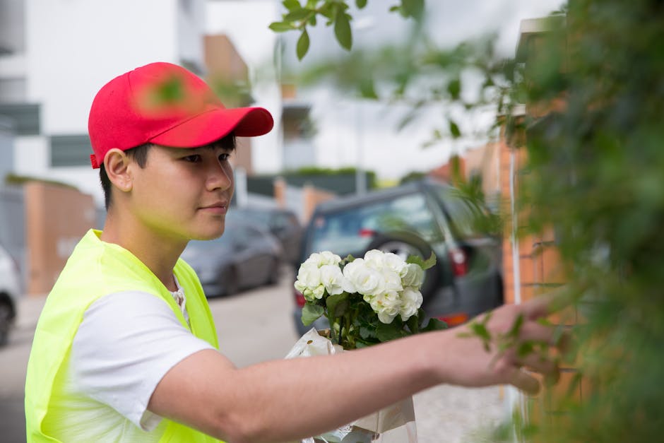 A young delivery person wearing a red cap and a high-visibility yellow vest is holding a bouquet of fresh white hydrangeas as they reach to place the floral gift against a leafy green hedge on a resid