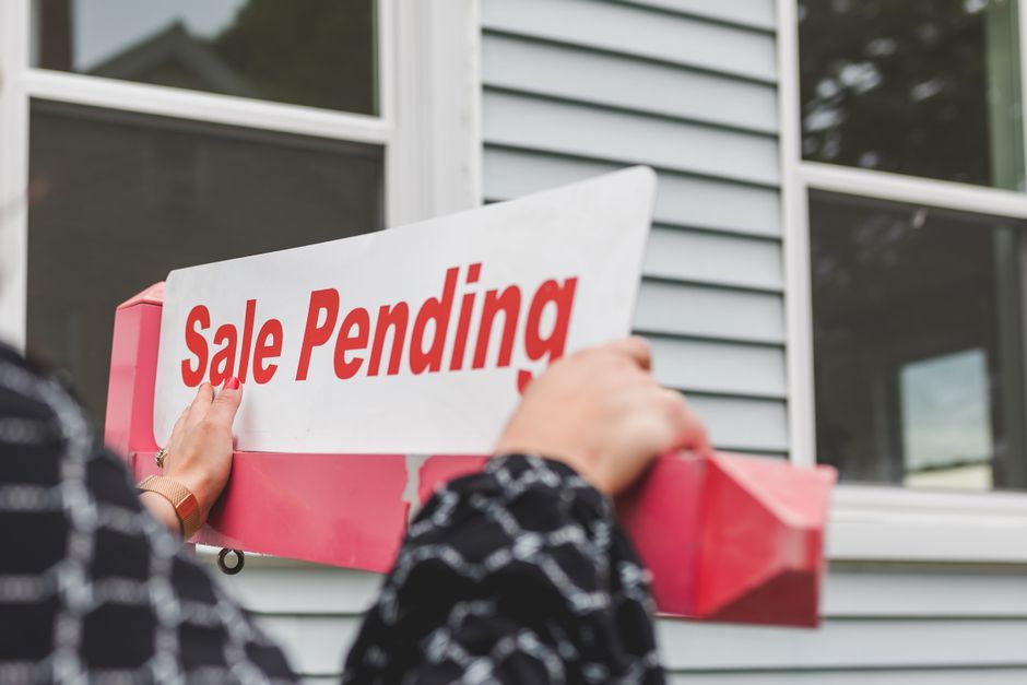 A person holding a rectangular pink box with a white sign that reads 'Sale Pending' in bold red letters. The individual is dressed in a black and white patterned outfit, and their hands are visible gr