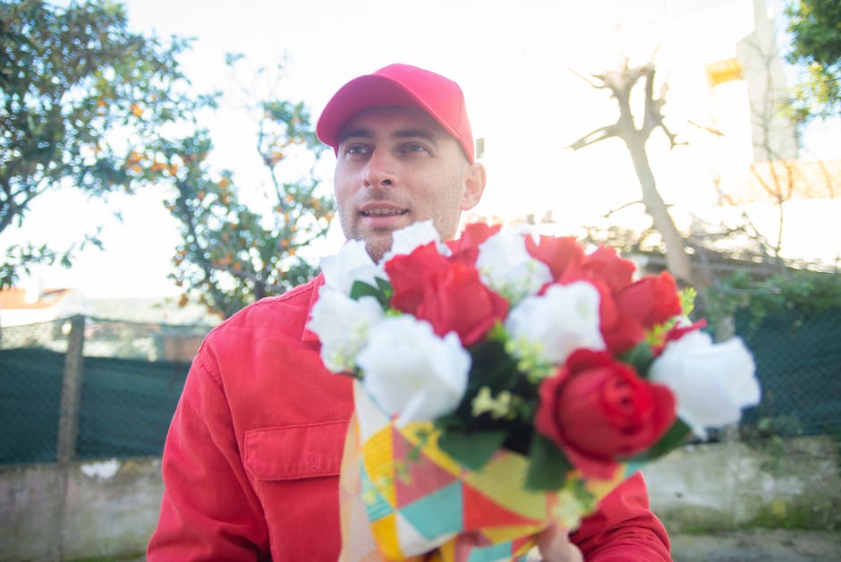 A man dressed in a red jacket and matching red cap holds a vibrant bouquet of fresh flowers towards the camera, with a mix of red, white, and green blooms. The bouquet is wrapped in colorful paper, an