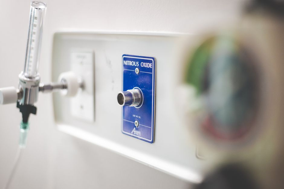 Close-up image of a medical oxygen supply outlet labeled 'Nitrous Oxide' mounted on a white wall, featuring a metallic valve socket and a connected green-tinted oxygen flow tube. The outlet is part of