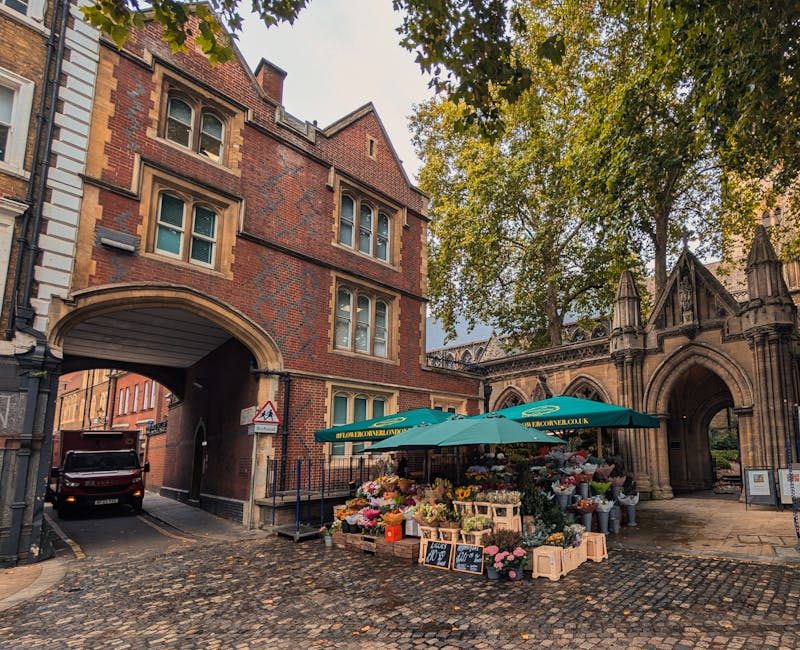 A flower stall set up on a cobbled street under a green canopy, displaying an assortment of fresh flowers including pink roses, white lilies, yellow tulips, and purple asters arranged in wooden crates