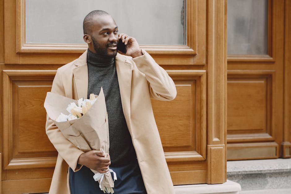 A man with dark skin, a neatly trimmed beard, and a friendly expression is standing outdoors in front of a wooden-paneled building, holding a bouquet of fresh white roses wrapped in brown craft paper.