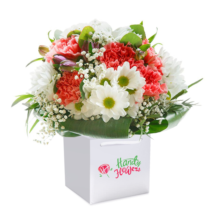 A radiant fusion of blossoms sits upright in an immaculate white cube-shaped gift bag, photographed against a seamless white background that accentuates the floral colours and crisp lines. The bouquet is dominated by sumptuous coral, soft peach, and vibrant red carnations, their ruffled petals forming a lush, tactile mosaic and infusing the display with the warmth and exuberance one finds along Thornton Heath's Greville Road in summer. Interspersed elegantly are clusters of snow-white daisies, each bloom shining with a distinct yellow-lime center that brings a sense of cheerful brightness and clarity. Wispy sprays of baby's breath introduce a light, airy texture, while a few gently closed buds edged in muted purples provide depth and intrigue-a subtle signal of new life and careful curation. The entire arrangement is ringed by masses of deep green foliage, the leaves' glossy surfaces and broad shapes enveloping the bouquet like a protective embrace. Sturdy white ribbon handles arch above the bag, promising easy delivery and transport to friends' doorsteps across Thornton Heath. The overall composition is balanced, rounded, and opulent, carefully crafted for those seeking to mark life's celebrations-be it a birthday, anniversary, or meaningful thank-you-with something truly memorable. Light falls delicately across the scene, illuminating the freshness of every petal and the skilled craftsmanship that brings together each element. This floral creation embodies the artistry and dedication of Thornton Heath's local florists, turning everyday moments into something extraordinary.