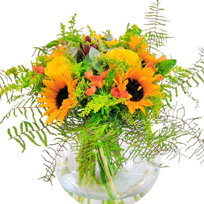 The first glance at this handcrafted bouquet from Thornton Heath brings an instant sensation of warmth and celebration - a visual echo of golden summer days. Towering sunflowers with their signature chocolate-brown centers and vibrant yellow-orange petals radiate positivity right from the heart of the arrangement. Intertwined, lush yellow roses spiral open with generous layers, their soft yellow tone recalling the delicate blooms scattered along the paths near Thornton Heath Recreation Ground. Scarlet Hypericum berries glisten among the foliage, smartly punctuating the palette with dashes of spirited red. Verdant greenery spills outward, with each feathery fern frond and spiky stem moving gracefully, adding a refreshing sense of motion that mimics the gentle flutter of leaves in the local parks. Airy solidago, laced with tiny yellow blossoms, nestles among the petals, while bluish eucalyptus leaves emerge at playful angles, infusing the bouquet with a breezy, natural scent. Contained within a sparkling round glass vase, the arrangement's green water-immersed stems are clearly visible, reinforcing the display's freshness and structural artistry. Standing as a thoughtful gift or an eye-catching centrepiece, this bouquet invites the openness and neighbourly cheer of Thornton Heath into any celebration - whether it's a milestone birthday, a new arrival, or simply a moment that calls for happiness made tangible.