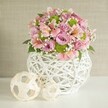Bathed in delicate natural light, this exuberant floral centerpiece captures the spirit of a Thornton Heath occasion-whether wedding, anniversary, or simply a thoughtful gesture for a loved one nearby. The arrangement is artistically composed with full bloomed pastel pink and lavender roses, their petals plush to the touch and visually striking. Interwoven among them, pink alstroemeria-known for their slender, trumpet blossoms and intricate striped interiors-contribute layers of gentle elegance. Scattered lime green button chrysanthemums add a playful touch, their spherical forms contrasting with the softness of tiny, floating flakes of baby's breath nestled throughout. Lush green leaves peek from beneath, anchoring the bouquet and suggesting freshness. The flowers rise from an eye-catching white vase, its open-lattice weave crafted from interlocked, slender strips, creating a blend of rustic inspiration and contemporary design. On the left, two decorative spheres, one large and one smaller, each constructed with pale, woven material, add visual interest and a sense of artisanal continuity. All of this sits atop a clean, white surface, against a beige-toned, textured background reminiscent of Thornton Heath's local character and welcoming homes. This scene reflects both artistry and warmth, a tangible representation of the care and dedication local florists bring to every arrangement entrusted to their hands.